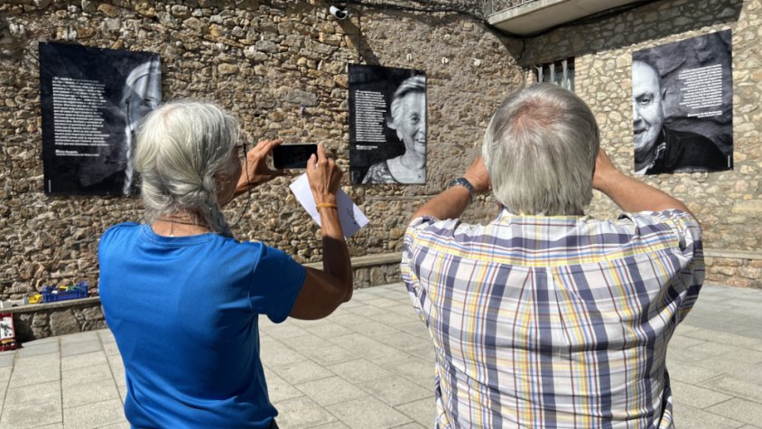 Dos veïns de Montellà fent fotografies a les lones que contenen retrats de gran format amb experiències de vida de la gent gran del municipi